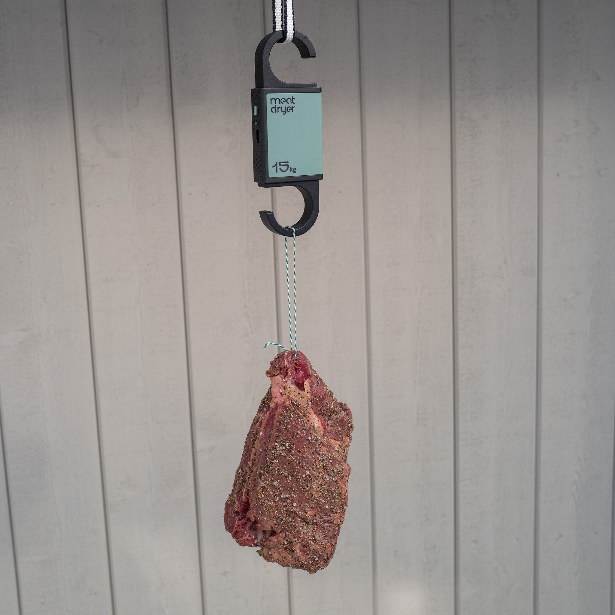 Biltong beef meat hanging from a green Meatdryer hook with a weight , temperature and humidity indicator against a grey background